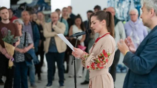 Fotografie einer jungen Frau die auf ein Blatt schaut und durch ein Mikrophon zu einem interessierten Publikum spricht. Rechts im Bild ist im Anschnitt ein Gebärdensprachdolmetscher zu sehen.