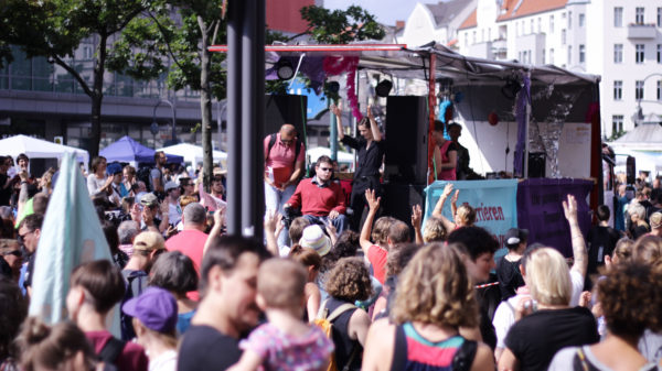 Urheberrecht Vivien Cahn. Foto eines geschmückten Umzugs-LKWs in einer Menschenmenge am Hermannplatz. Es ist sonnig und die Stimmung wirkt ausgelassen. Auf dem LKW sind Menschen, ein junger Mensch im Rollstuhl hält eine Rede.