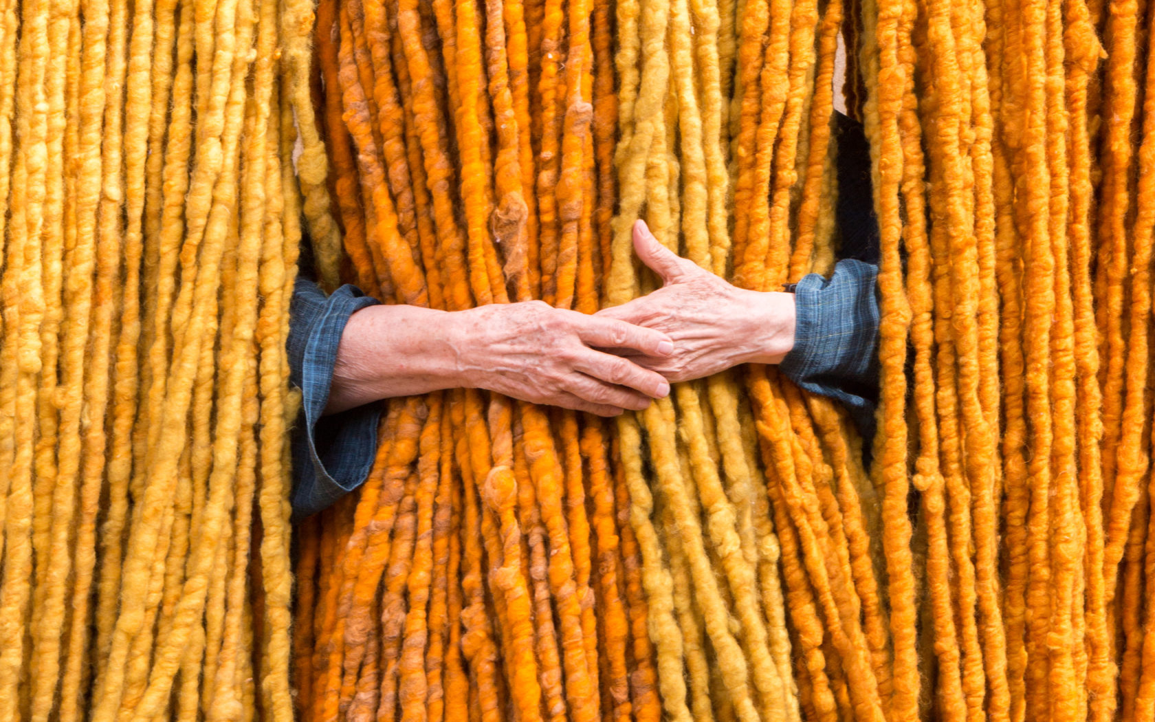 Von der Decke hängen dicke, gelb-orange Stoff-Fäden, die von hinten von einer älteren Frau umarmt werden. Man sieht nur ihre Hände. 