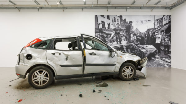 Vor einem Schwarz-Weiß-Bild, das eine protestierende Menschenmenge zeigt, steht ein silbernes Auto, das stark beschädigt ist. Die Fenster des Fahrzeugs sind zerschlagen, die Glassplitter liegen auf dem Boden. Mehrere Beulen verunstalten die Karosserie. Im Bild sind viele Menschen zu sehen, die protestieren, während einige Kinder auf einem anderen Auto herumspringen.